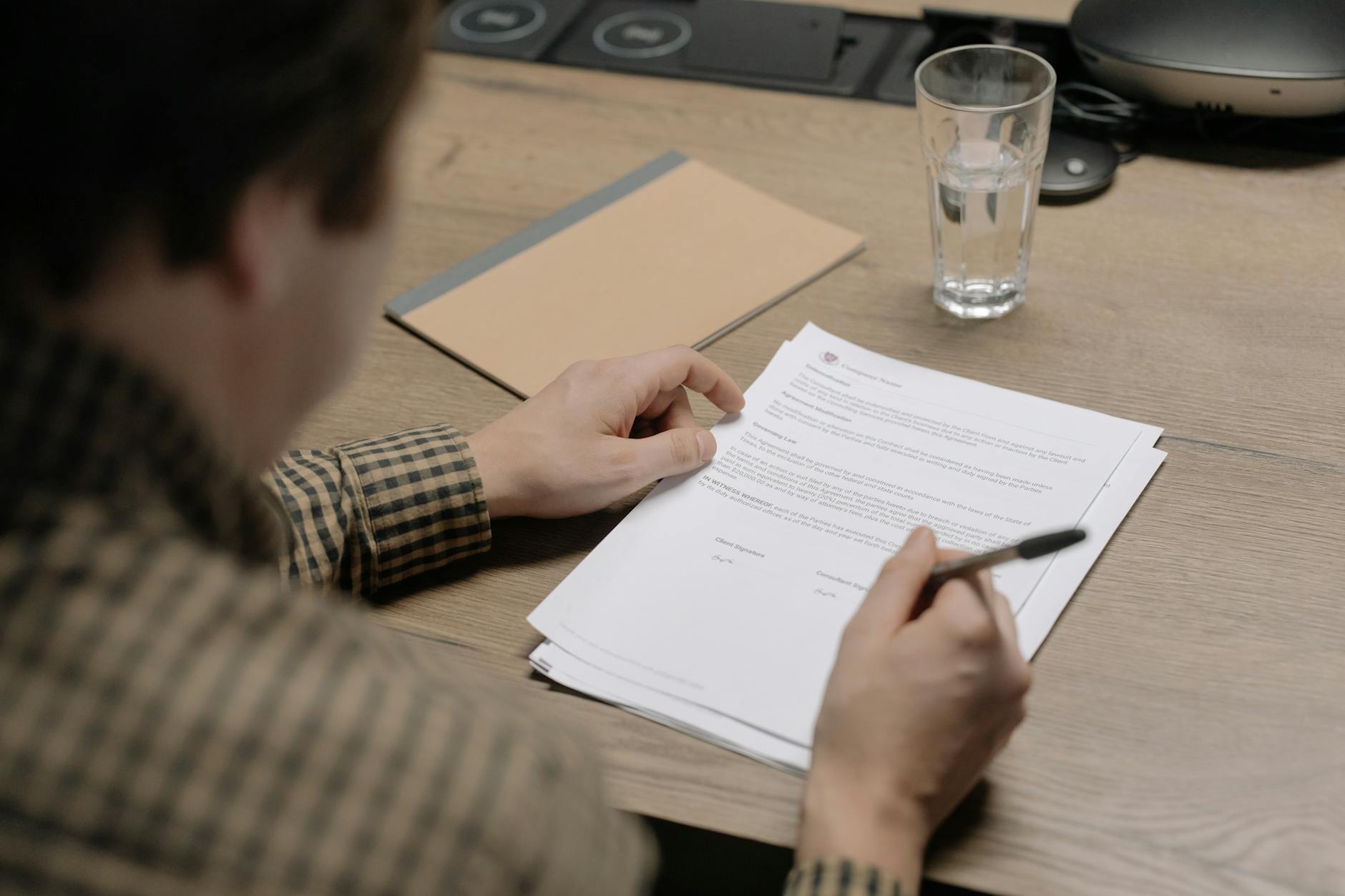 A businessman is reviewing a contract while holding a pen at a desk with a glass of water nearby.
