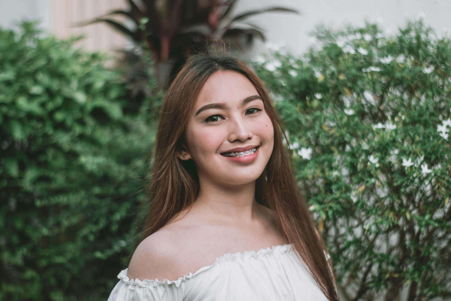 Portrait of a young woman with braces smiling outdoors against green foliage.