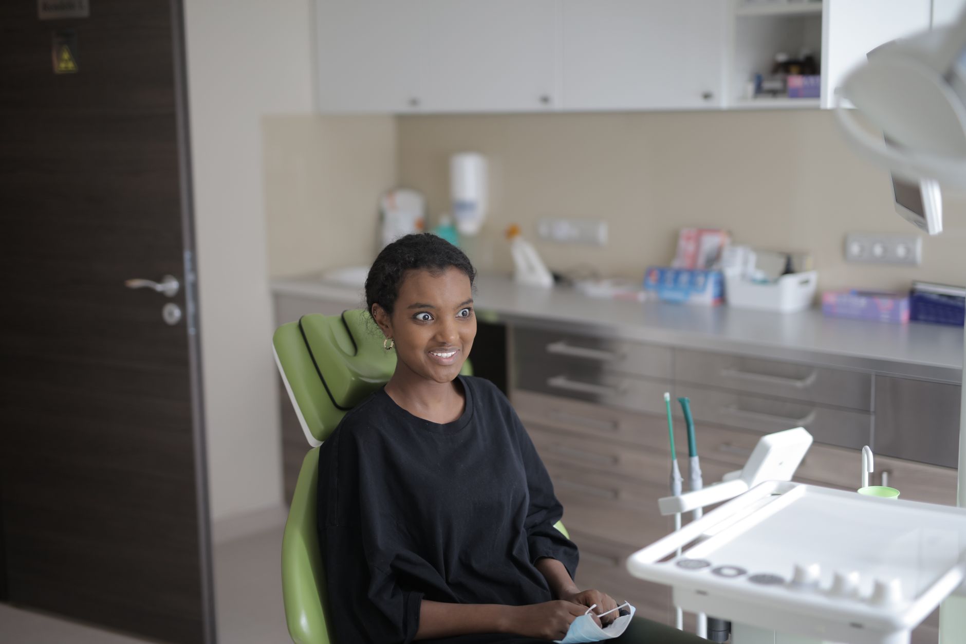 Surprised young ethnic female client in black casual clothes sitting in dental chair and looking away before treating teeth in modern clinic