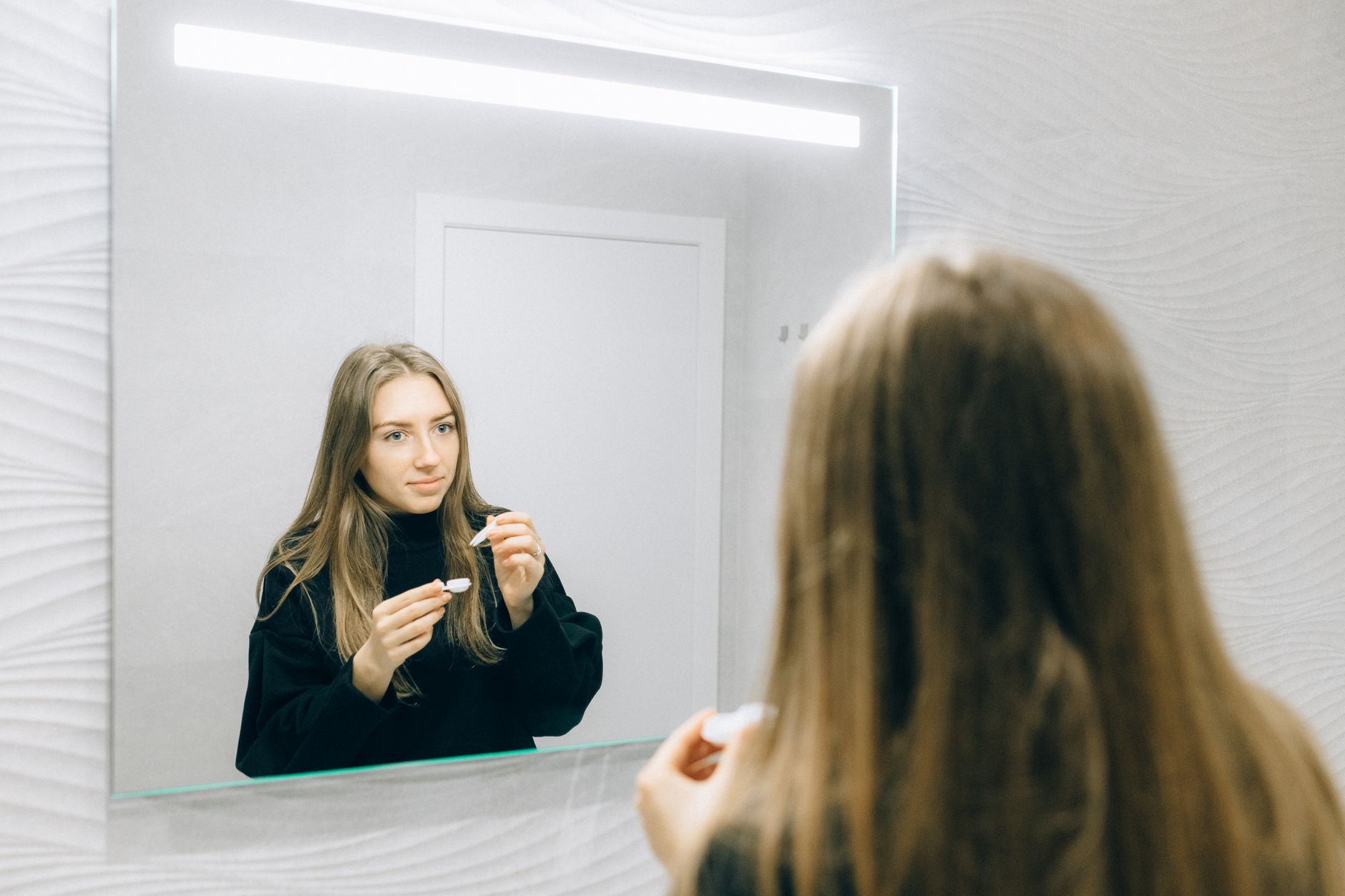 Young woman examining colored contact lenses in mirror, comparing cosmetic lens options for eye color change