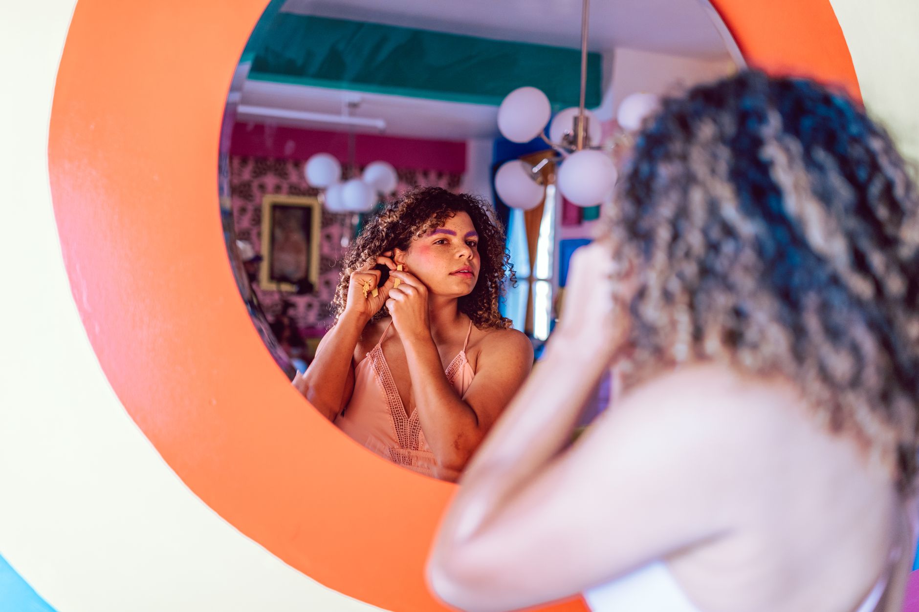 A woman with curly hair adjusts her earring while looking into a colorful, circular mirror indoors.