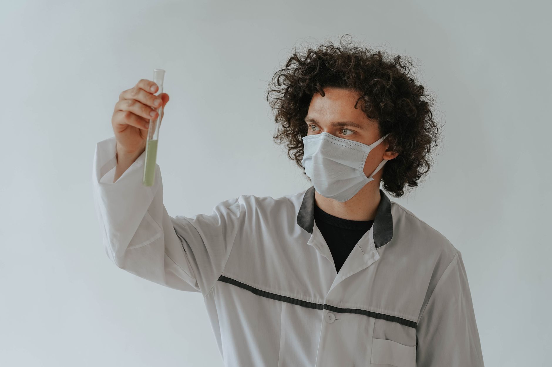 A scientist in a lab coat examining a test tube while wearing a face mask.