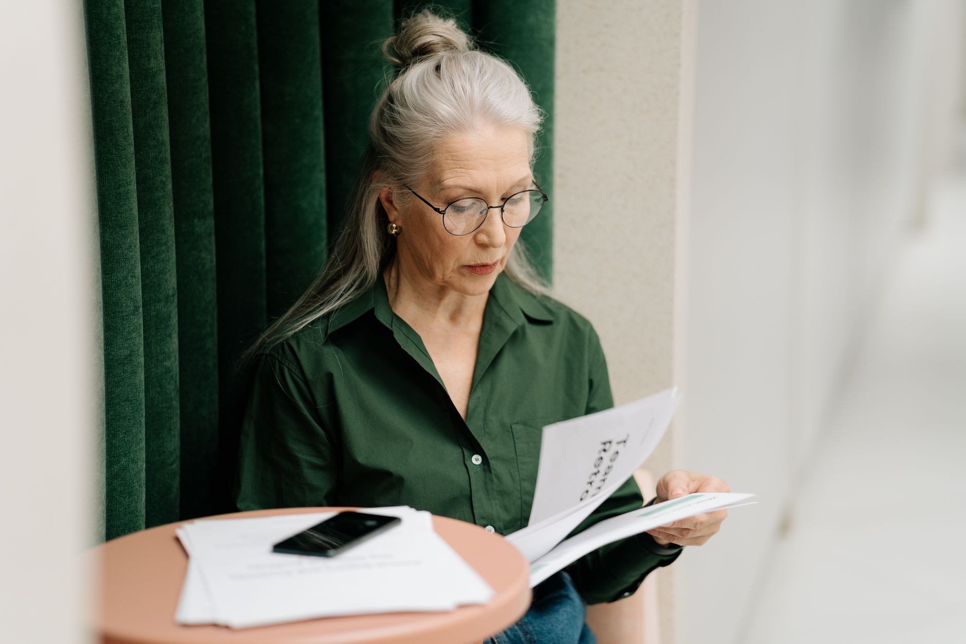 Elderly woman with gray hair reading papers at a small table indoors, focused and thoughtful.