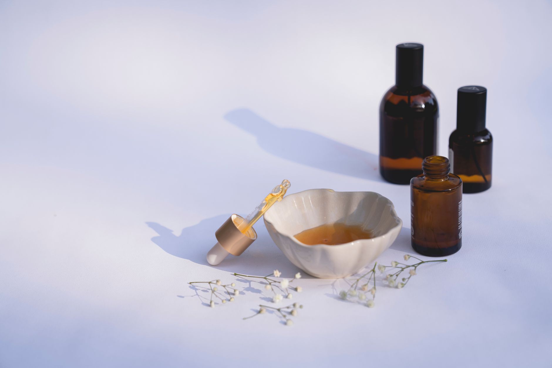 Elegant minimalistic display of amber glass bottles, ceramic bowl, dropper, and flowers on white background.