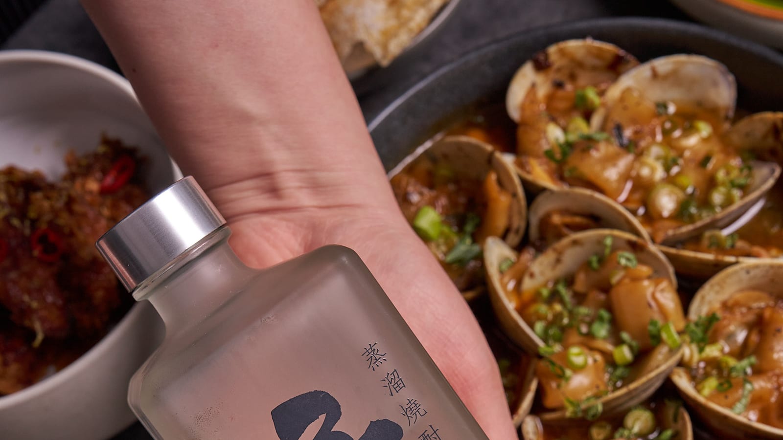 Hand holding a bottle of Korean soju above a table of clams and rice at 8282