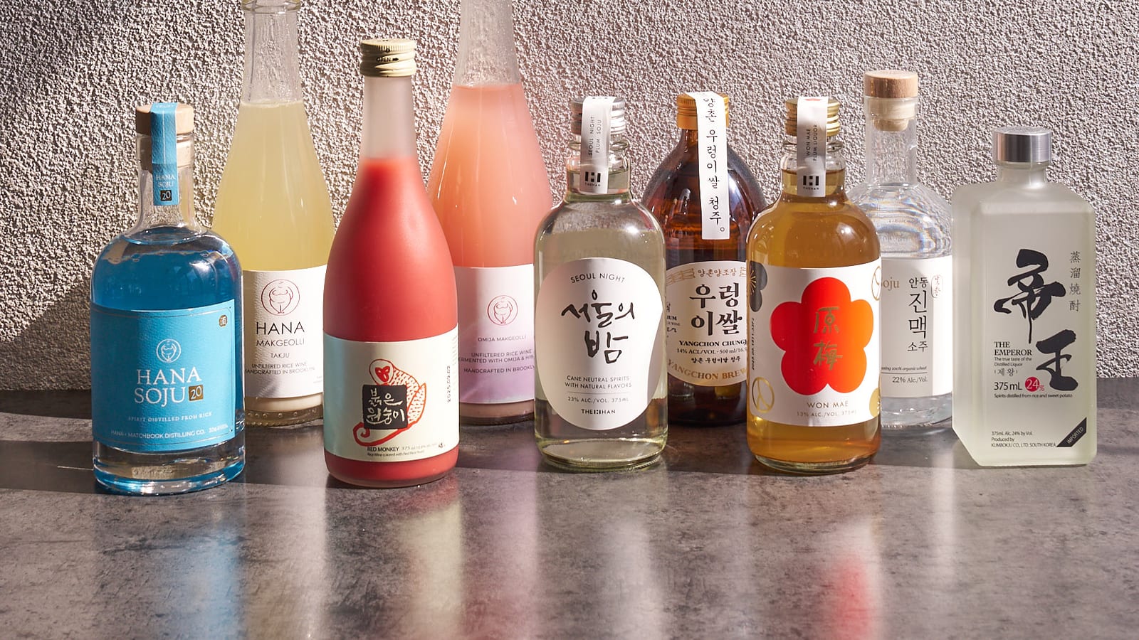 Lineup of Korean spirit and makgeolli bottles against a grey wall